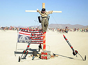 Decorative missles mounted on the ground point at the face of a gagged effigy tied to a cross. An inverted flag tagged with anarchist graffiti is propped up nearby.