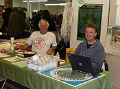Two men sit at a showroom table, surrounded by swag.