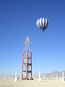 A hot air balloon hovers behind the Burning Man effigy, which stands upon a four-story, open rectagular framework.